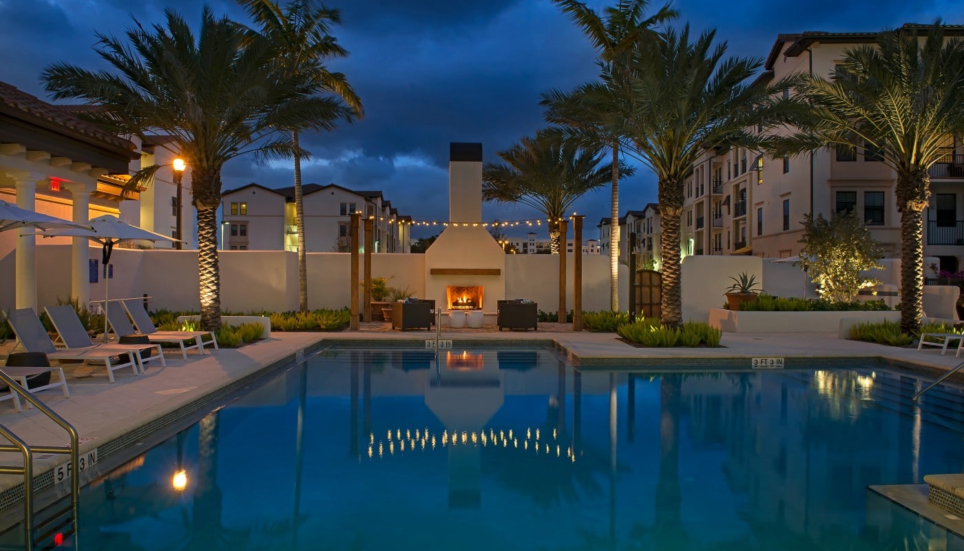 light house point resort style pool at night with chaise lounge chairs, umbrellas, palm frees and fireplace - jefferson apartment group