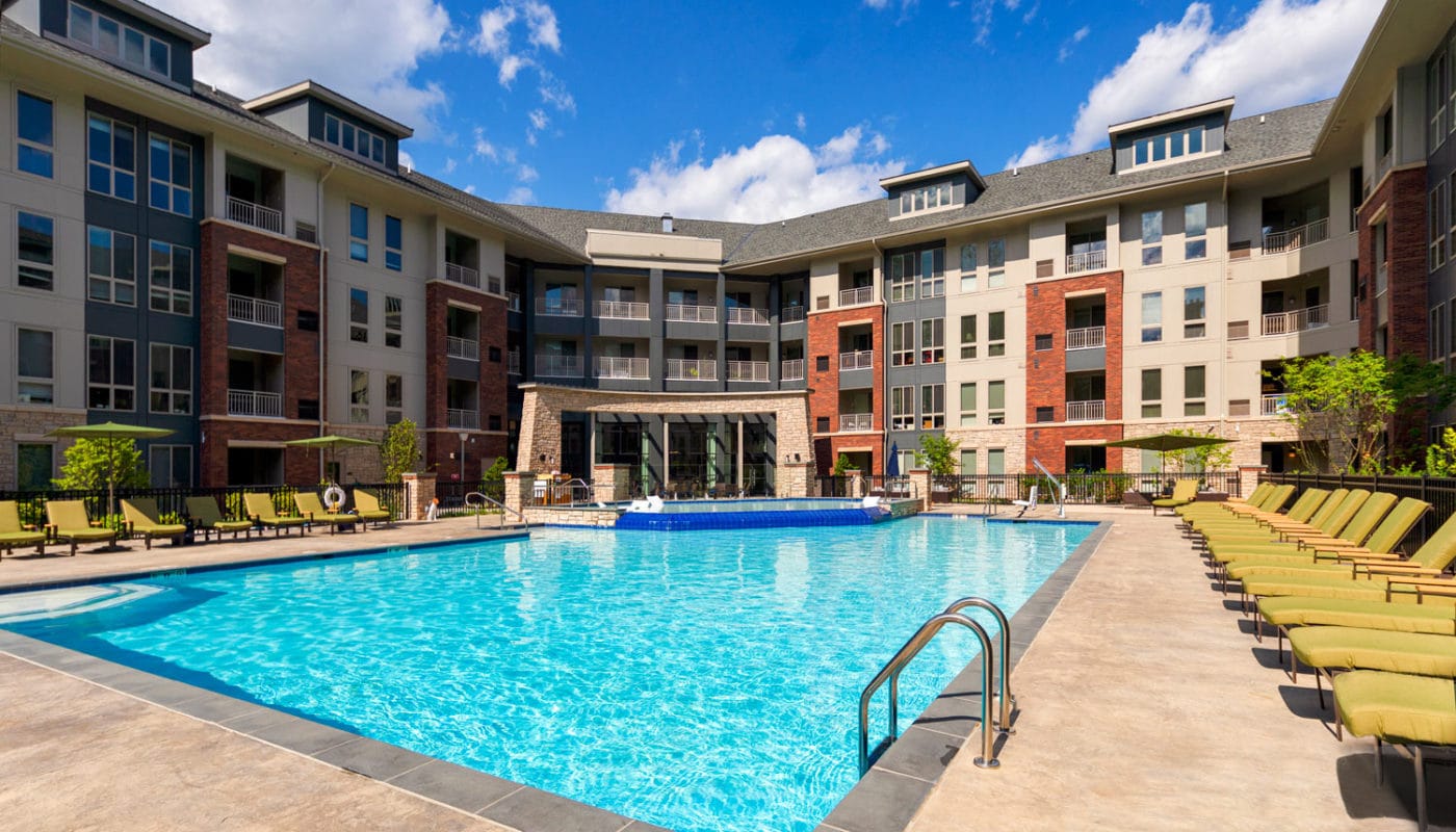 maybrook resort style pool with chaise lounge chairs, umbrellas, greenery, and view of apartment building in the background - jefferson apartment group