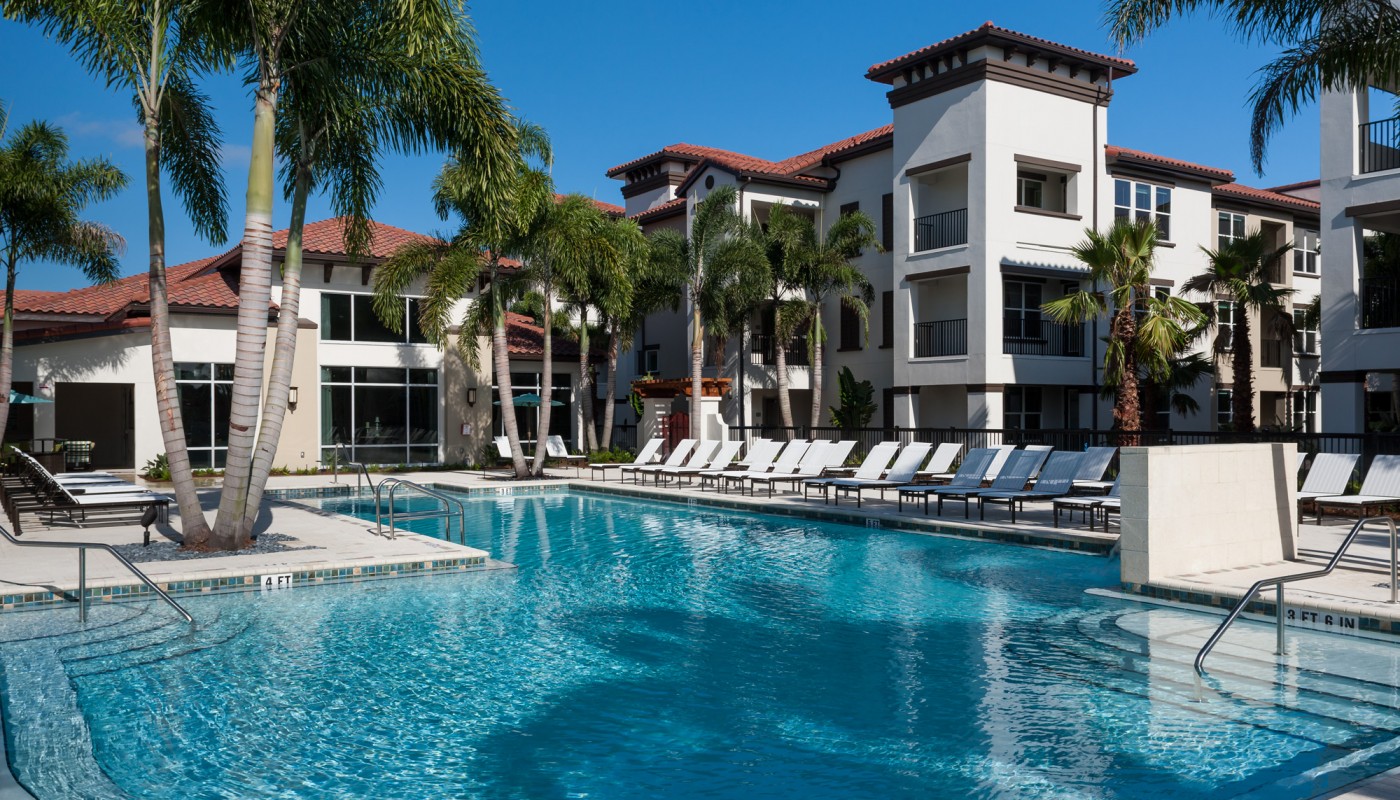 westshore resort style pool with chaise lounge chairs, pergola, palm trees and view of apartment building and wooded area in the background - jefferson apartment group