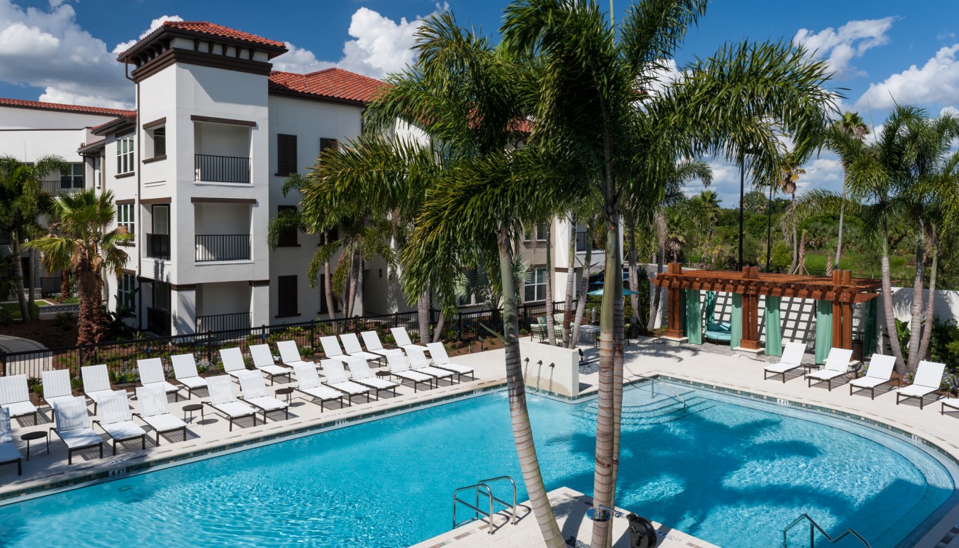 westshore resort style pool with chaise lounge chairs, pergola, palm trees and view of apartment building and wooded area in the background - jefferson apartment group