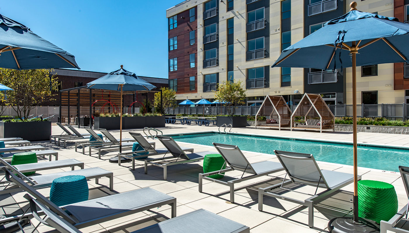 j malden center resort style pool with chaise lounge chairs, umbrellas, cocktail tables and view of apartment building in the background - jefferson apartment group